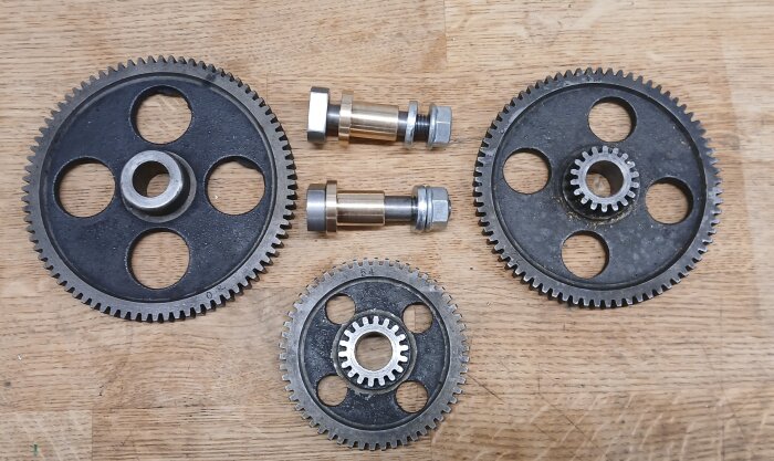 Två nya axlar med bussningar och tre kugghjul, varav ett märkt "54", på ett trädgolv.