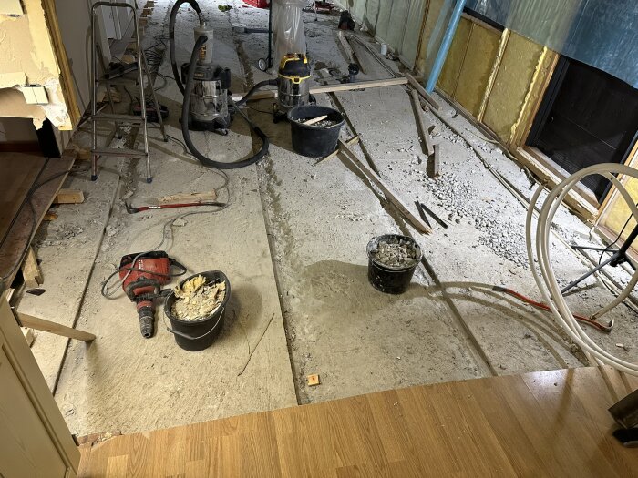 Renoveringsprojekt med bilad golv för avloppsrör, verktyg och slangar synliga. Väggar med isolering och plast, stege och dammsugare i bakgrunden.