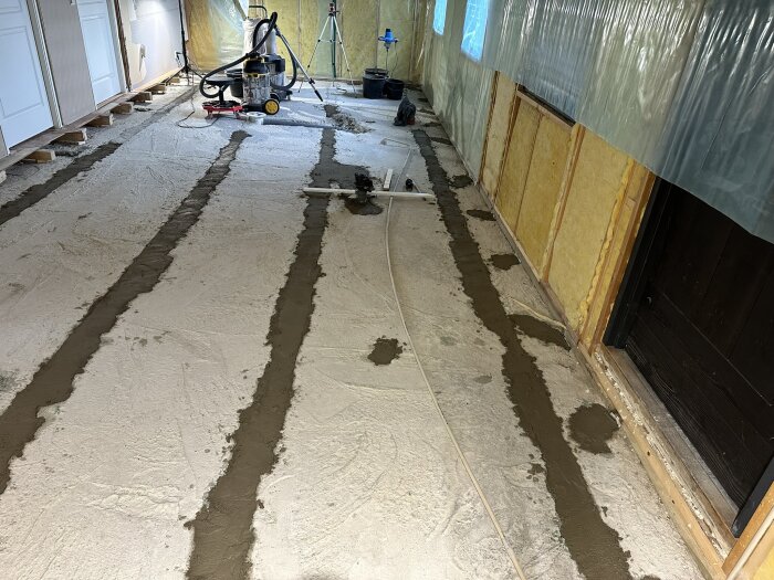 Översiktsbild av byggarbetsplats inomhus med nygjutna betongrännor på golvet och plastad vägg, verktyg och dammsugare i bakgrunden.