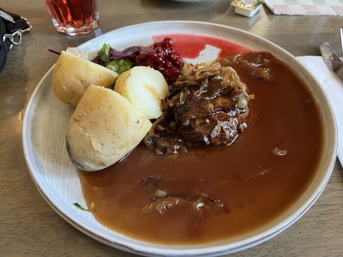 Tallrik med pannbiff, sås, potatis, sylt och sallad på ett träbord med en röd dryck i ett glas bredvid.