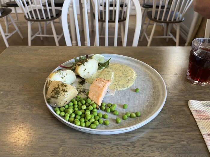 Tallrik med lax, gräddsås, kokt potatis och gröna ärtor på ett bord i en restaurang.