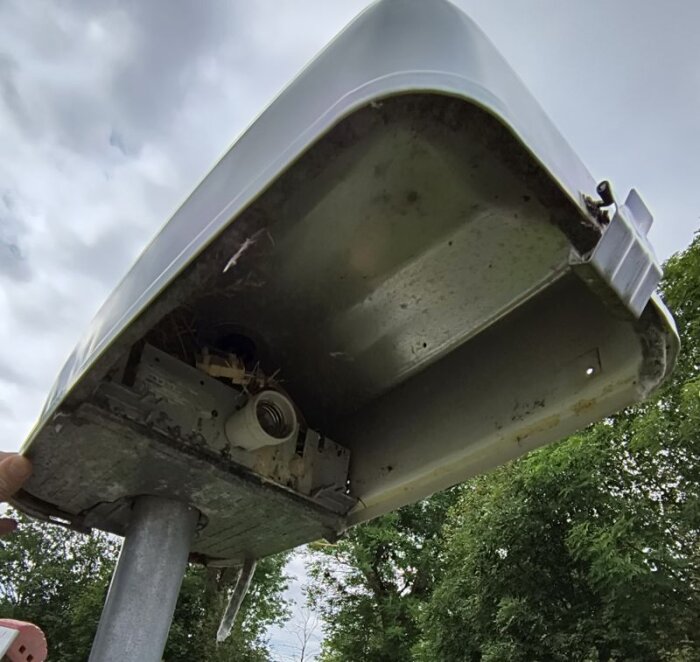Gatubelysning öppnad, visar insidan med fågelbo och rusten, samt en tom lampsockel, belägen ovanpå en metallstolpe och omgiven av träd.