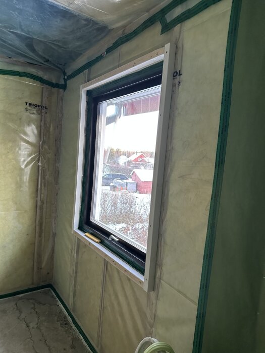 Rum under renovering med plastväggar, ett fönster och delar av golvet synliga. Utomhus ses snötäckta träd och röda hus.
