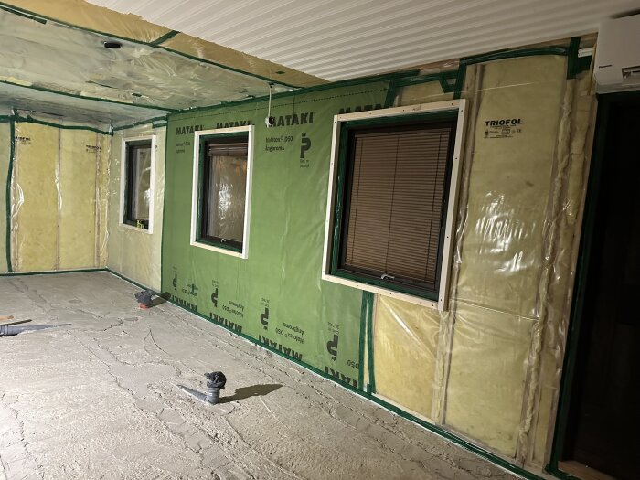 Renoveringsbild av rum med väggar täckta av isolering och plast, två fönster, synliga rör på betonggolv; arbete pågår, november 2024.