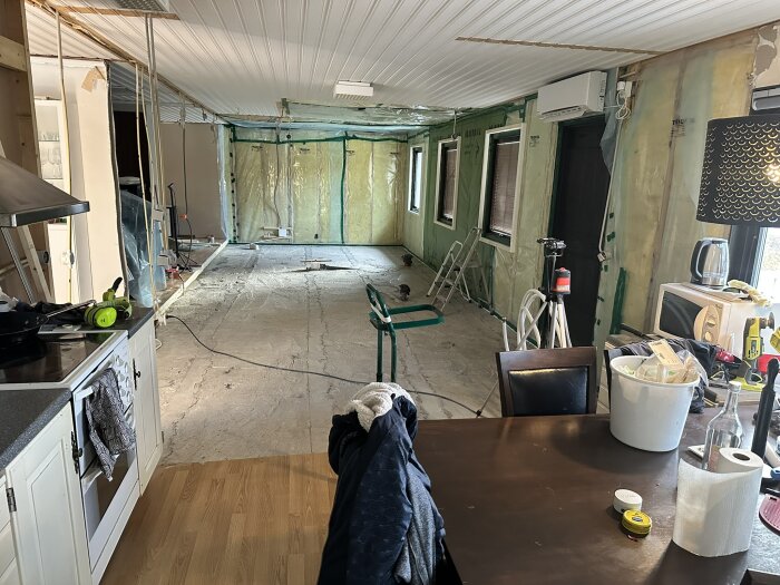 Översiktsbild av en pågående renovering; köksutrustning i förgrunden, rivna väggar och plastade ytor i bakgrunden. Byggmaterial och verktyg syns.