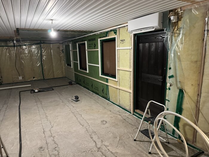 Rum under renovering med isolerade väggar, plastfolie och en dörr. Stege och byggmaterial finns på det ojämna betonggolvet.