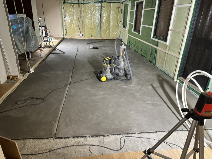 Flytspacklat golv i ett rum under renovering med verktyg och utrustning, inklusive dammsugare och stativ, samt väggar med grön isolering.