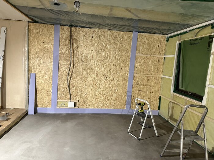 Ett rum under renovering med OSB-skivor på väggarna, lila våtrumsgips runt om. Stegstegar och en borrmaskin syns på golvet.