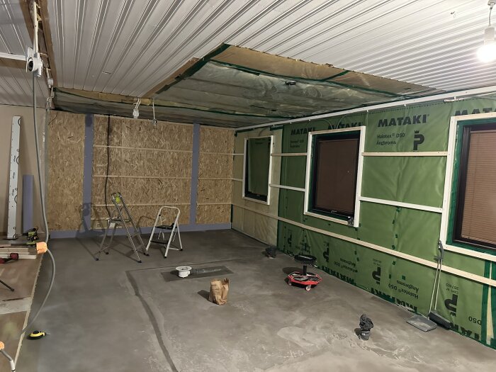 Renoveringsprojekt: rum med gröna väggar av isolering, fönster, stegar, verktyg och oskimpat betonggolv, redo för installation av golv- och väggmaterial.