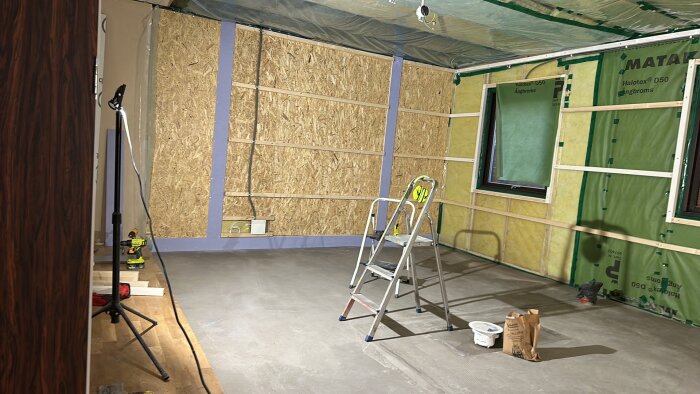 Ett rum under renovering med nylagd flytspacklad golvyta, reglade väggar och en stege. Plast och gipsskivor syns på väggarna.