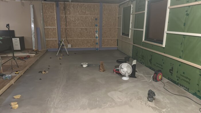 Renoveringsrum med reglar, grön plastfolie, och betonggolv. Verktyg och fläkt syns på golvet. Väggar delvis gipsade i våtrum.