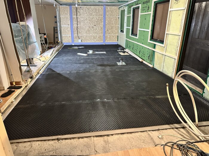 Installationsskikt av Jape Ventgolv matta på ett golv under montering, med oisolerade väggar i bakgrunden. Slangar och verktyg syns i rummet.