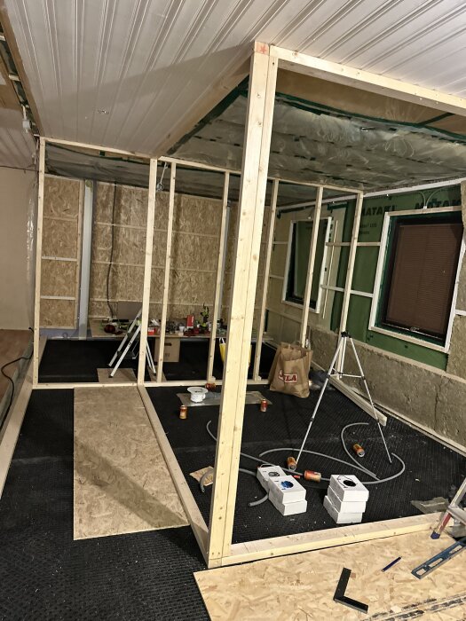 Rum under renovering med träramar för väggar och tak, isolering, verktyg och byggmaterial synliga på golvet.
