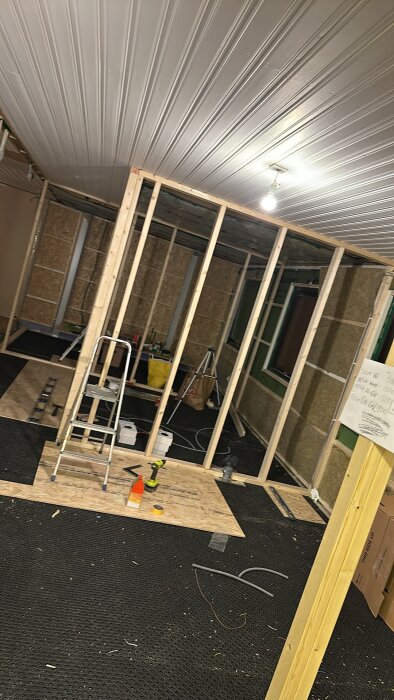 Bygge av innerväggar med träreglar, en stege står lutad mot väggen och olika verktyg och material syns på golvet.
