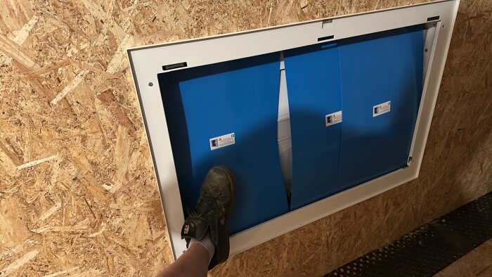En vit fördelarskåp med blå dörrar monterat i en OSB-vägg, foten av en person trycker mot skåpet.