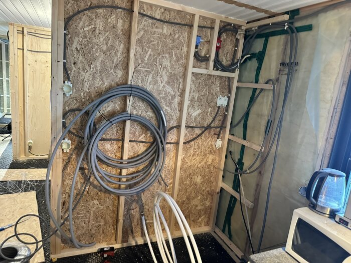 Vägg med oisolerade installationsrör och eluttag monterade på träpanel, kablarna är oordnat upphängda och golvet har temporära skyddsskivor.