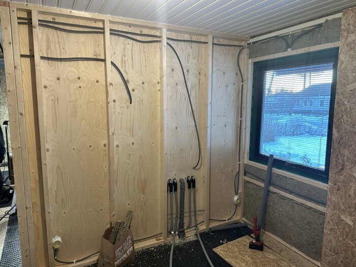 Vägg med regling och synliga rör för installation av golvvärme, ett fönster till höger med persienner. Verktyg och material på golvet.