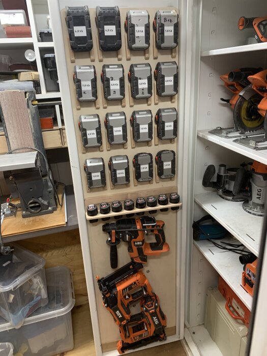 Väggmonterad laddstation med 18 batterier och två verktyg i en verkstadshylla, inklusive borrmaskin och spikpistol.