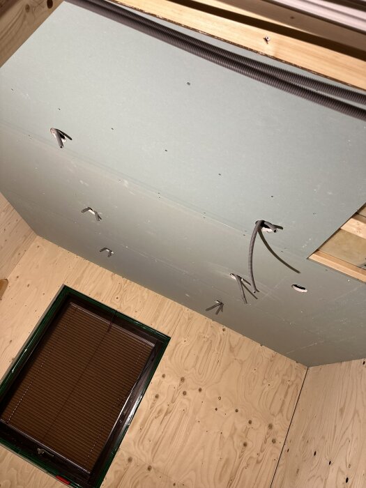 Tak i badrum under konstruktion, gips med utskurna hål för infällda spotlights och synliga kablar, plywoodvägg med litet fönster med persienner.
