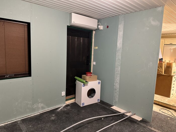 Renoverat rum med gröna gipsväggar, inplastad yttervägg och tak med infällda spottar, samt byggmaterial på golvet.