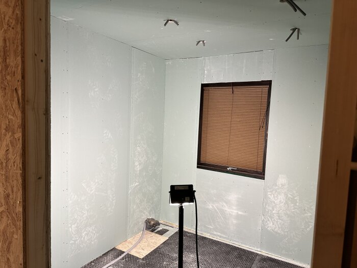 Badrum med gröna gipsskivor och svart golv, fönster med persienn, LED-lampa och synliga takfästen för infällda spottar.