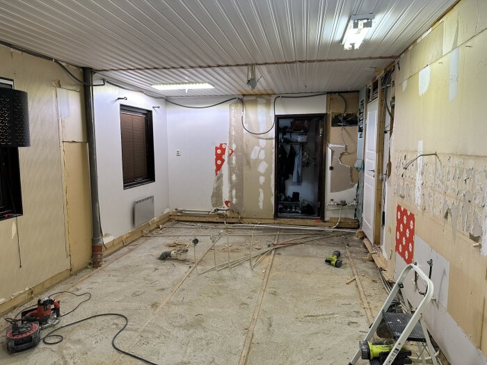 Renoveringsprojekt i hus under rivning med synliga rör på golv, avskalade väggar och taklampor, verktyg och stege synliga i rummet.