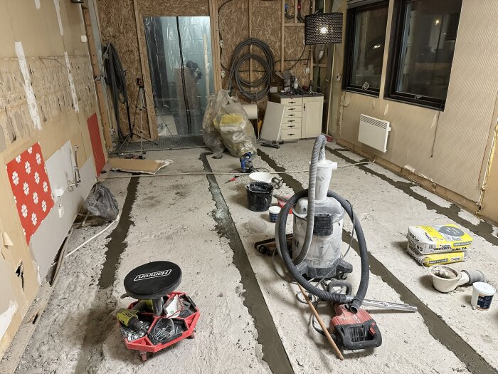 Renoveringsarbete i kök med verktyg och material; betonggolv under bearbetning, dammsugare central i bilden, plastdörr för dammskydd i bakgrunden.