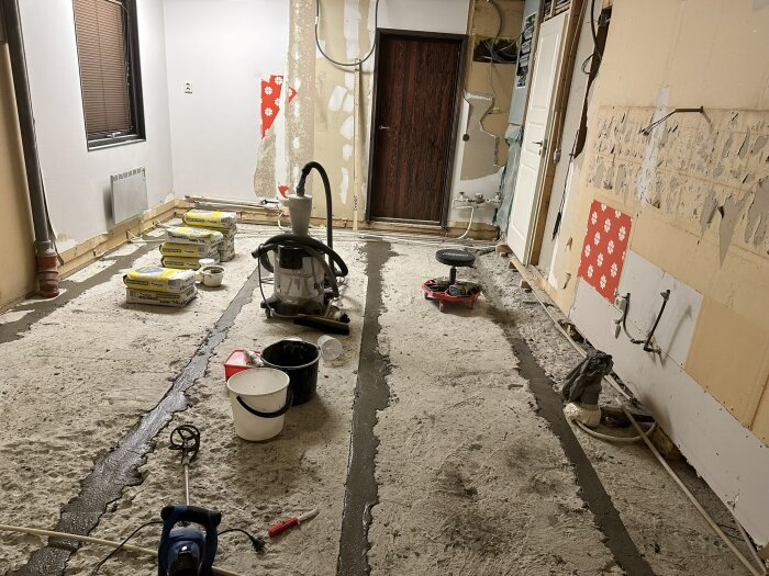 Renoveringsprojekt i kök med rivna väggar och golv, byggmaterial staplat och verktyg synliga i rummet, fläckar av flytspackel på golvet.