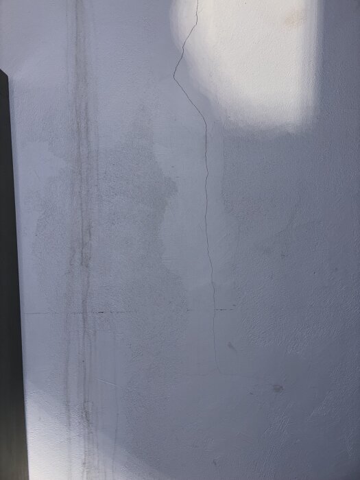 Spricka i vit putsad innervägg med smal vertikal spricka och äldre målningsförsök. Ljus och skugga reflekteras på väggen.