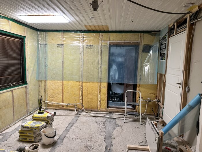 Renoverat rum med isolerade väggar och plasttäckt öppning, byggmaterial och verktyg synliga på golvet, ingen inredning.