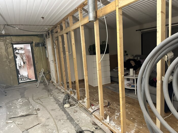 Renovering av interiör visar delvis riven vägg mellan vardagsrum och kök, med blottade träreglar och byggmaterial på golvet.