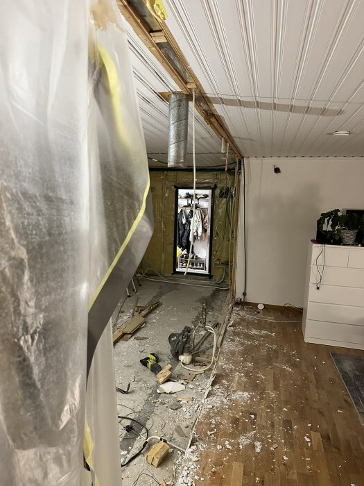 Renovering av rum med riven vägg mellan kök och vardagsrum, byggmaterial och verktyg ligger på golvet, taket exponerat och plast täcker delar av rummet.