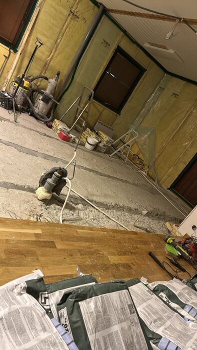 Renoveringsarbete med blottat golv, isolerade väggar, verktyg och byggmaterial inomhus. En dammsugare och rör ses på golvet.