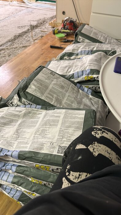 Säckar med flytspackel staplade på ett golv, verktyg synliga i bakgrunden, del av ett renoveringsprojekt i ett vardagsrum.