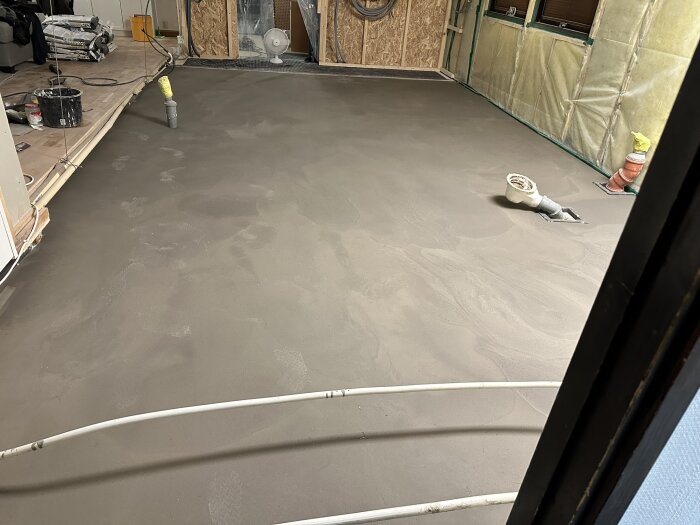 Flytspacklat golv i ett rum under renovering med rörinstallationer synliga och byggmaterial staplat i bakgrunden.