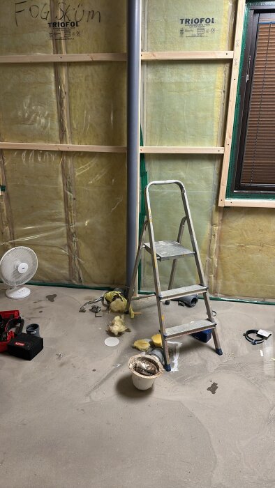 Stege och verktyg på ett golv i ett rum under renovering, med isoleringsmaterial på väggen bakom.