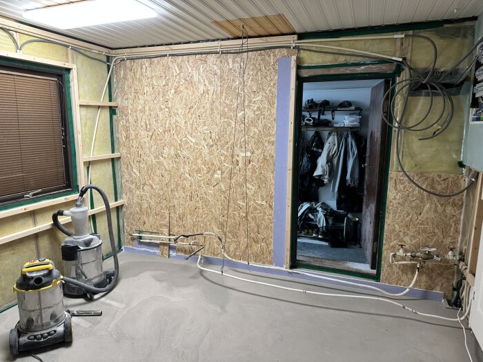 Rum under renovering med osb-väggar, isolering, rör, och dammsugare. Dörr öppnar till rum med kläder och skor; golv förberett för flytspackling.