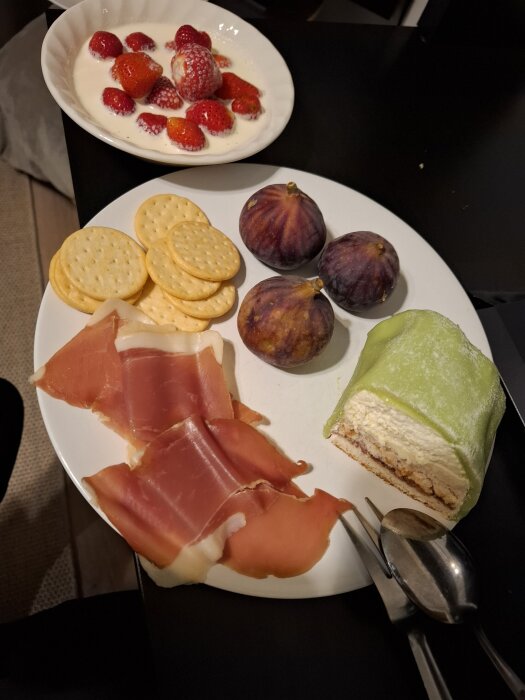 Tallrik med prinsesstårta, fikon, lufttorkad skinka och salta kex. I bakgrunden en skål med jordgubbar och grädde.