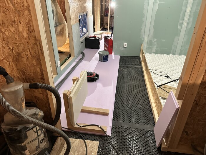 Byggarbetsplats med isolering och verktyg; rosa cellplast och LK Systemskiva syns, samt en dammsugare och annan utrustning i ett rum med gröna väggar.