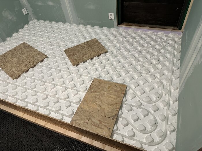 Golvvärmeslangar installerade i ett rum, med vita cellplastskivor och två plywoodbitar ovanpå.