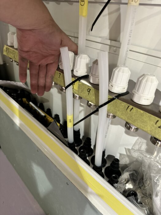 Hand placerar rör vid installationsskåp för värmesystem. Numrerade rör och kopplingar synliga, verktyg och material i närheten.