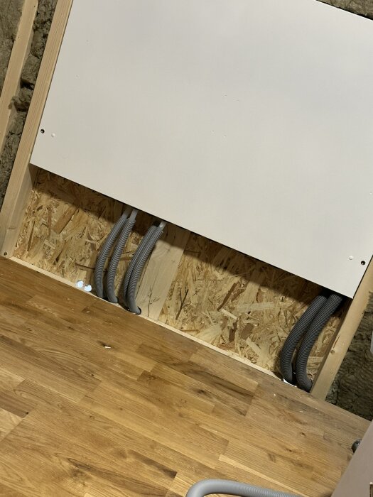 Isolerade väggar och golv med sex grå slangar installerade under en vit panel, synlig trästruktur bakom.