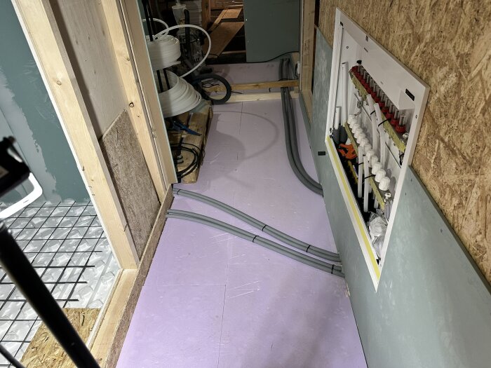 Installationsområde med golvvärmesystem, rör och reglar på trävägg. Kontrollpanel för golvvärmeslangar synlig på väggen.