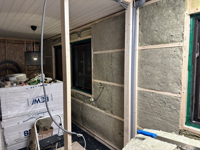 Byggarbetsplats inomhus med isolering på väggar, verktyg och material runtomkring. Taklampa hänger över staplade kartonger.