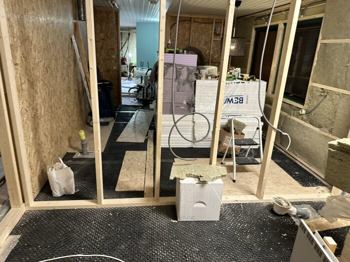 Utrymme under renovering med träreglar, isolering, och byggmaterial runtomkring; en stege står framför vit isoleringsskiva.