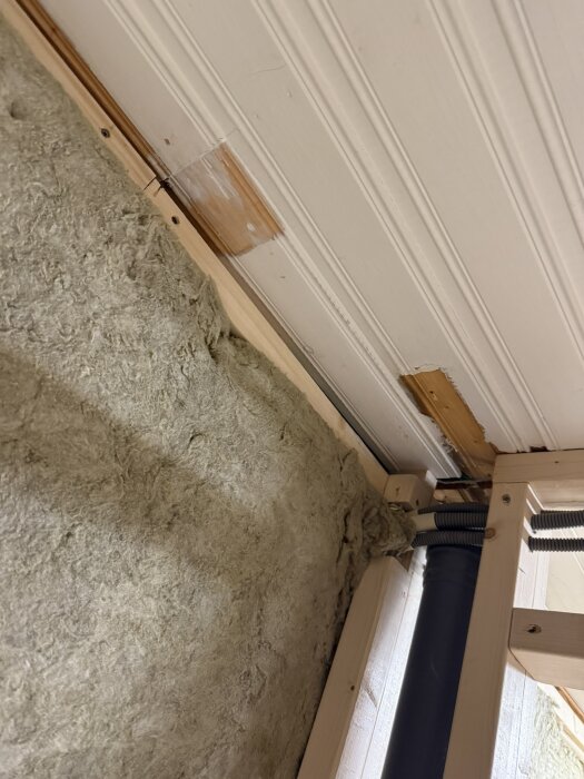 Isolering och reglar i byggprojekt, visar vägg med isoleringsmaterial, träreglar och rördragning i hörnet av ett rum med vitmålat tak.