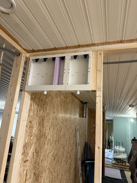 Monterat skåp för bergvärmepump i en träkonstruktion med synlig rördragning, sett från ett rum under renovering med träväggar och tak.