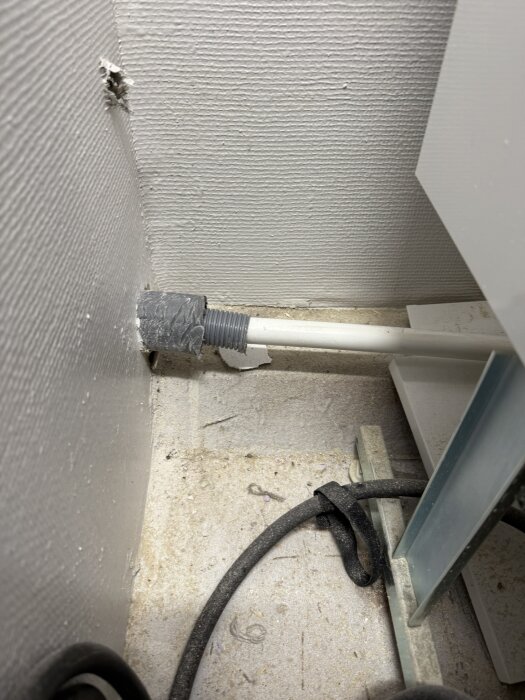 Vägghörn med installerad grå slang, synlig tejp och svart kabel på ett dammigt golv.