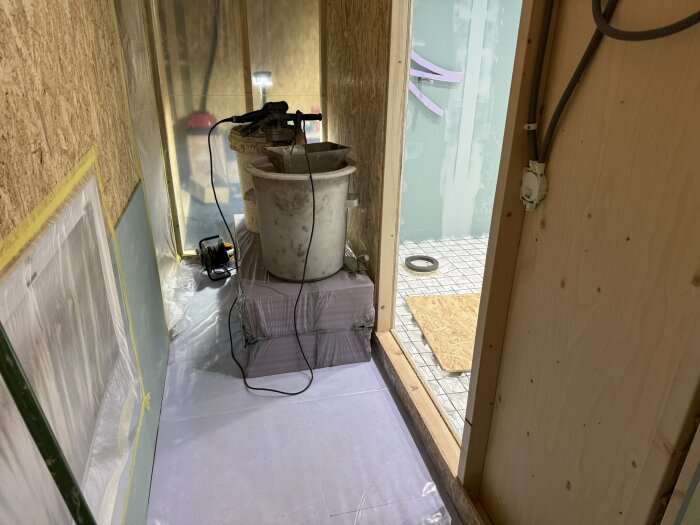 Byggarbetsplats med blandningsutrustning och slangrullar på ett täckt golv, del av pågående renoveringsprojekt i ett hus.