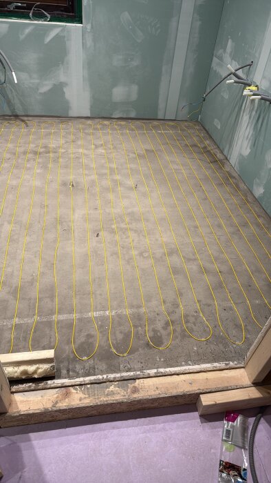 Elektrisk golvvärmeslinga på betonggolv i ett rum med gröna väggar under renovering. Slingorna är jämnt fördelade över ytan.